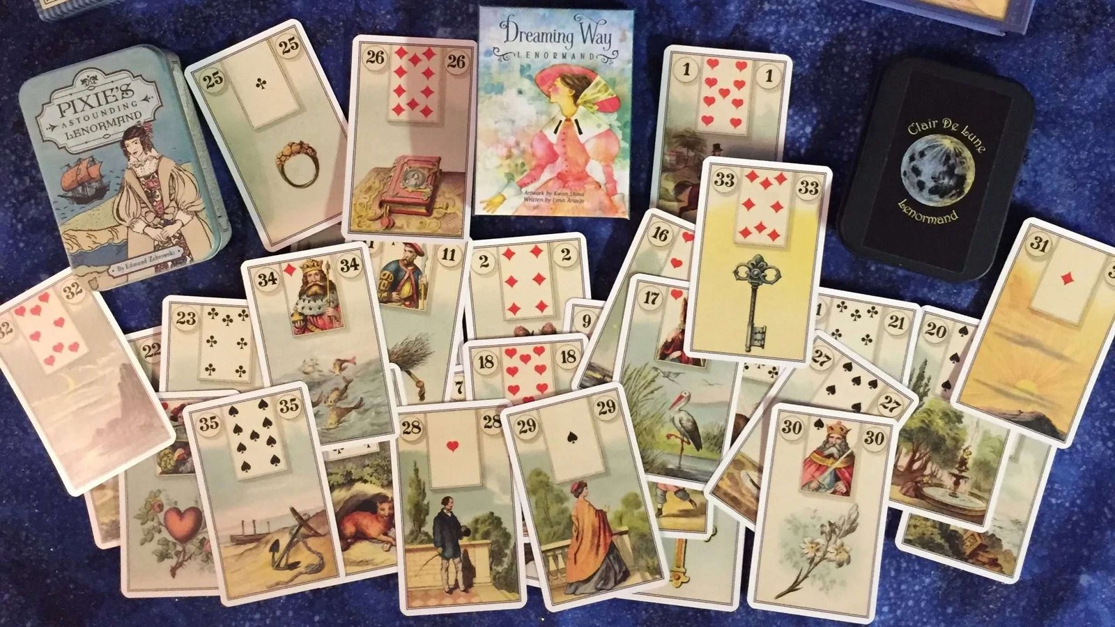 Cartas de Petit Lenormand espalhadas sobre um pano azul-escuro, ao lado de três baralhos em caixa.